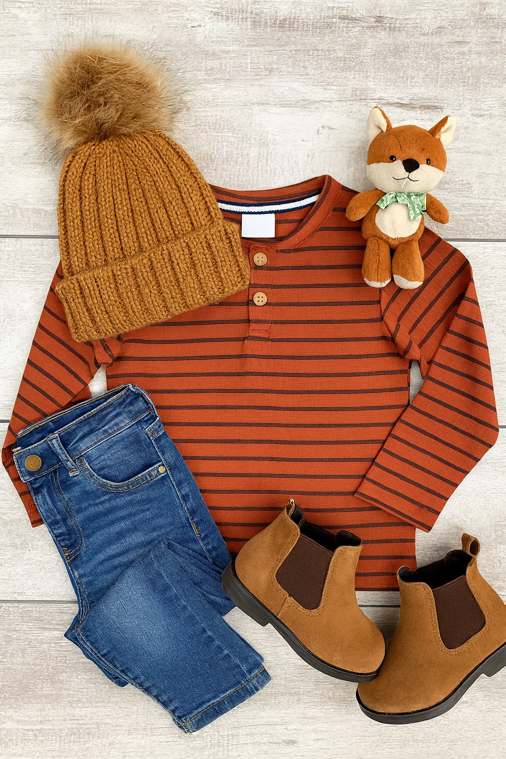 Children's outfit with striped shirt, jeans, boots, beanie, and plush fox toy on a wooden surface.